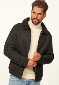 Schwarze Kunstwildlederjacke mit einem flauschigen Kragen, gerippten Bündchen und Saum. Mit Reißverschlussverschluss, getragen über einem cremefarbenen Rollkragenpullover und hellen Jeans.