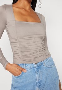 Långärmad, fyrkantig topp i ljus taupe-tyg med samlad textur och figurnära passform, ihop med ljusblå denimshorts.