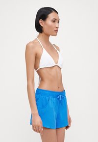 Balta bikini viršus su trikampio formos puodeliais ir plonais dirželiais, derinamas su ryškiai mėlynomis maudymosi šortais, turinčiais elastingą juosmenį ir šonines kišenes.