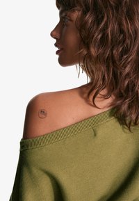 Olivově zelený svetr se spadlými rameny z měkkého úpletu. Má žebrovaný výstřih a malý tetování na rameni. Textura působí příjemně a útulně.