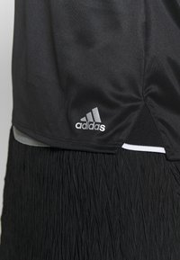 T-shirt de sport Adidas noir avec un logo sur le côté inférieur, porté sur une jupe ou un pantalon texturé noir, montrant les détails du tissu et des coutures.