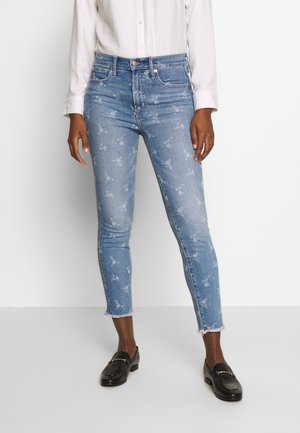 Vrouw draagt lichtblauwe skinny jeans met witte bloemenprint, rafelige zoom, zwarte loafers en een wit overhemd met knoopsluiting.