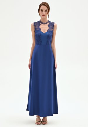 Donna in piedi che indossa un abito senza maniche di un blu reale con dettagli in pizzo sul corpetto e sulle spalle, abbinato a sandali dorati con punta aperta, su sfondo bianco.
