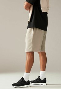 Beige Shorts mit Seitentaschen, kombiniert mit schwarzen Sneakers und weißen Knöchelsocken, vor einem neutralen Hintergrund.