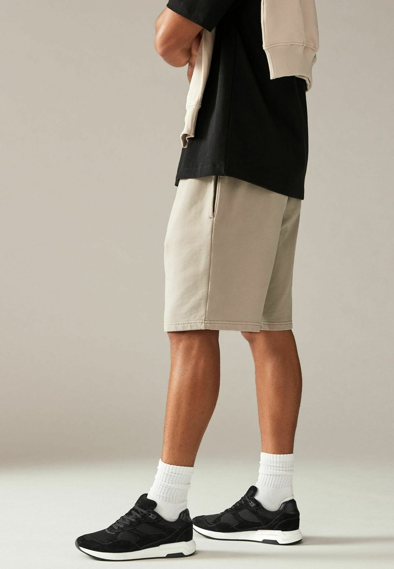 Beige shorts med sidelommer, kombineret med sorte sneakers og hvide ankelsokker, mod en neutral baggrund.