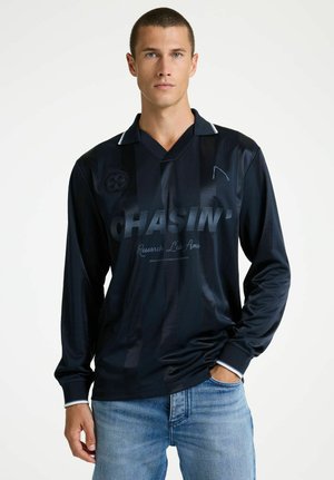 Muž v čiernej dlhorukávovej košeli s nápisom "CHASIN'" a svetlomodrými džínsami, stojaci s jednou rukou v vrecku na bielom pozadí.