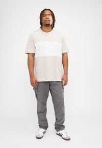 Beige t-shirt met een witte horizontale streep met reliëflogo, gecombineerd met grijze cargobroeken en witte sneakers met zwarte accenten.