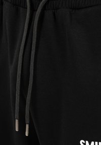 Gros plan sur un pantalon de jogging noir avec une ceinture à cordon de serrage et un texte blanc imprimé partiellement visible près de la poche.