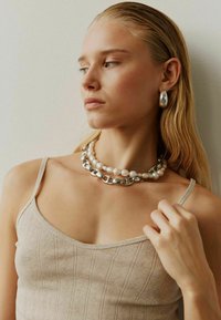 Bež pleteni top s tanke naramenice, slojevit sa bisernim i srebrnim lancima. Srebrne ovalne naušnice upotpunjuju izgled.