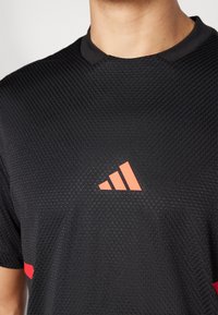 Čierne športové tričko s textúrovanou látkou, ktoré má červený akcentový prúžok a červené trojuholníkové logo adidas na hrudi.