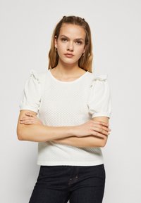 Top blanco de manga corta con mangas abullonadas, tela texturizada con pequeños agujeros, escote redondo y diseño corto. Combinado con jeans de mezclilla oscuros.