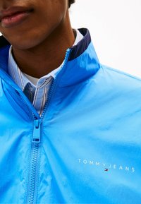Blauwe lichtejas met een hoge kraag, voorzien van een ritssluiting aan de voorkant en het logo "TOMMY JEANS". Het materiaal heeft een gladde textuur en een glanzende afwerking.