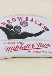 White fabric patch with red text "THROWBACKS" and "Mitchell & Ness." Features a black silhouette of a football player throwing a ball.