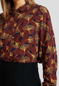 Chemise à manches longues bleu marine avec un motif de feuilles de ginkgo rouges et jaunes. Dotée d'une patte de boutonnage et d'un col. Tissu texturé.