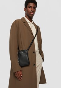 Bolsa de couro preta crossbody com fecho, usada sobre uma camisa bege e um casaco trench castanho claro. A bolsa apresenta um logótipo discreto e uma alça.