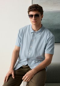 Helles blaues Kurzarmhemd mit strukturiertem, geripptem Design, mit Kragen und Knöpfen. Kombiniert mit braunen Hosen und Sonnenbrille.