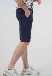 Shorts bleu marine en mélange de coton, longueur genou, avec des poches latérales et une étiquette au dos, portés avec des baskets blanches volumineuses.