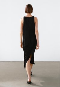 Robe noire sans manches avec une coupe ajustée et une fente sur le côté ; dotée d'un col haut et d'une fermeture éclair discrète au dos. Texture de tissu lisse.