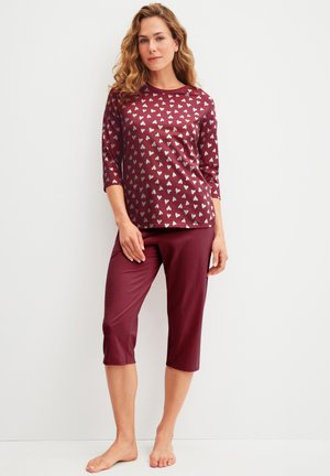Femme debout pieds nus, portant un haut à manches longues bordeaux avec motif de cœur et un pantalon court assorti sur fond blanc.