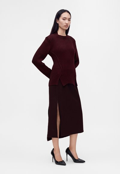 Femme debout portant un pull côtelé bordeaux foncé et une jupe midi assortie fendue à l'avant, associée à des chaussures à talons noirs.