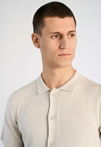 Ljusbeige polo-tröja i stickad kvalitet, med krage, korta ärmar och tre knappar fram med en diskret logotyp broderad på bröstet.