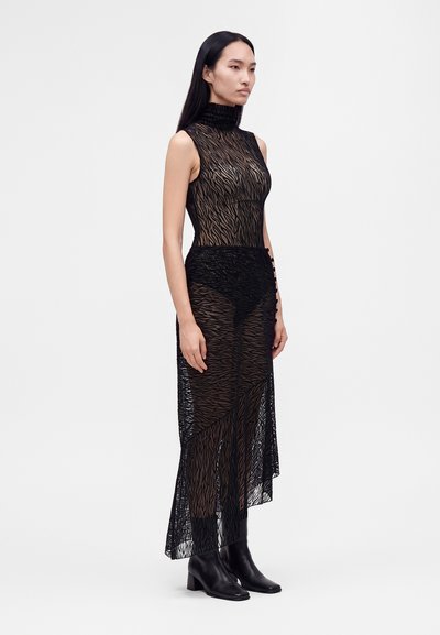 Femme portant une robe noire sans manches en tissu transparent avec un col montant et un motif zèbre, associée à des bottines noires, debout devant un fond uni.