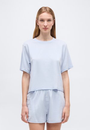 Ung kvinde med glat lysebrunt hår iført en lyseblå kortærmet skjorte og matchende løse shorts mod en ensfarvet baggrund.