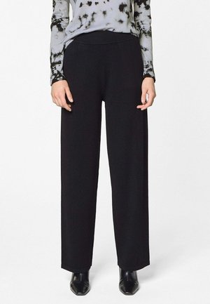 Femme portant un pantalon noir à jambes droites, des bottes noires à bout pointu, et un haut gris à manches longues avec des motifs tie-dye noirs.