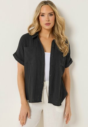 Blondynka w czarnej krótka koszula z guzikami na białym topie i białych spodniach, stojąca na tle prostej jasnej ściany.