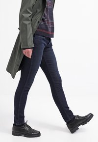 Mörkblå skinny jeans med en lätt stretch, kombinerade med en grön trench coat och svarta snörskor. Slät tygtextur, åtsittande design.