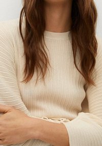 Femme aux longs cheveux bruns portant un pull en tricot côtelé crème, les bras croisés doucement sur le corps.