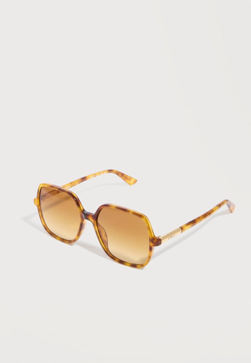 Lunettes de soleil carrées en écaille de tortue avec des verres dégradés marron et des branches fines, inclinées légèrement vers la droite sur fond blanc.