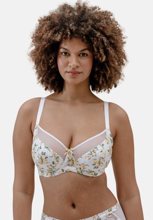 Frau mit lockigem Haar, die einen weißen BH mit gelben Zitronen und transparenten Mesh-Einsätzen trägt, blickt nach vorn vor einem einfarbigen Hintergrund.