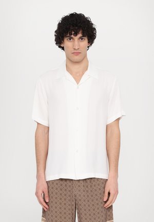 Camisa blanca de manga corta con cierre de botones y tejido suave, combinada con pantalones marrones de patrón geométrico.