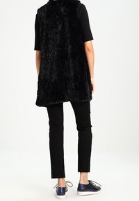 Svart fuskpälsväst med en texturerad yta, kombinerad med svarta byxor och blå snörskor. Slät tygkvalitet och knälång design.