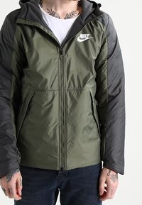 Nike huvjacka med en olivgrön kropp och mörkgrå ärmar, dragkedjestängning och logotyp på vänster bröst, med en mjuk textur.