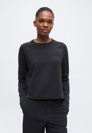 Femme aux cheveux courts portant une chemise noire texturée à manches longues et un pantalon noir, debout les mains dans les poches devant un fond clair.