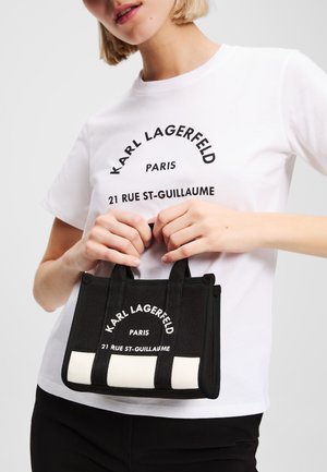 Borsa in tessuto nera con accenti bianchi, caratterizzata da testo in grassetto e due manici corti. Indossata da una persona in una t-shirt grafica bianca.