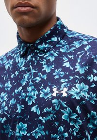 Κοντινό πλάνο ενός άνδρα που φοράει μπλε πουκάμισο με γιακά, με ανοιχτό μπλε λουλουδάτα σχέδια και λευκό λογότυπο Under Armour στο στήθος.