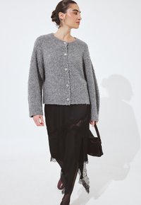 Pelēks fakturēts kardigans ar pārmērīgiem piedurknēm un piecu pogu aizdari, uzlikts virs melnas svārku kleitas ar mežģīņu apdari un melnas rokassomas.