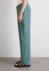 Lichtgroene, loszittende linnen broek met een elastische tailleband en trekkoorddetail, gecombineerd met zwarte sandalen. Zijaanzicht.