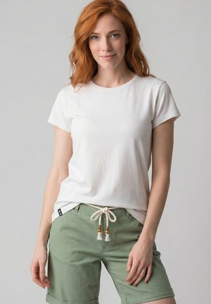 Rothaarige Frau, die ein weißes kurzärmliges T-Shirt und grüne knielange Shorts mit einem weißen Seilgürtel trägt, steht vor einem grauen Hintergrund.