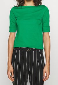 Grön kortärmad topp med rund halsringning, gjord av ett slätt tyg. Bärs med svart och vit randig högt skuren byxor.