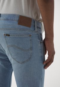 Light blue denim jeans with a brown leather brand patch. Features straight cut, back pockets with stitching detail, and subtle texture.