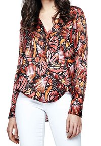 Femme portant un chemisier à motifs de papillons manches longues, associé à un pantalon blanc, se tenant avec les mains détendues.