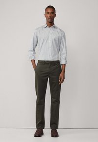 Camicia a righe blu chiaro e bianca con bottoni, abbinata a pantaloni verde oliva scuri. Il modello è in piedi dritto, con le mani in tasca e indossa scarpe marroni.