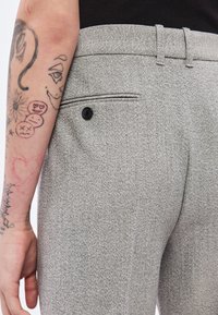 Halli tekstuuriga püksid, millel on kohandatud lõige, tagatasku musta nupu aktsendiga. Lähedalt vaadatuna tekstiili detailid ja käsivarte tätoveeringud.