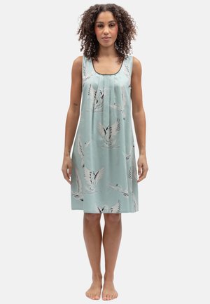 Vrouw met krullend haar die een mouwloze lichtblauwe jurk met een witte zwaan-print draagt, staat blootsvoets tegen een effen achtergrond.