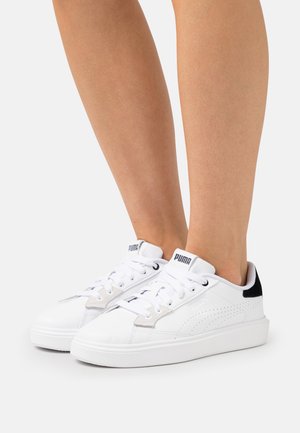 White sneakers with perforated sides, flat laces, a textured sole, and a black heel accent. Features a logo on the tongue.