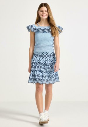 Ung pige smilende, iført en lyseblå kjole med mørkeblå blomsterbroderi og hvide sneakers, stående mod en ensfarvet baggrund.